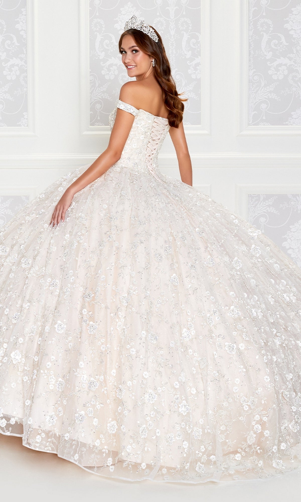 A woman in a sparkling Princesa Glow-in-the-Dark Quince Dress PR12266 with an off-the-shoulder neckline, full skirt, and tiara stands before an ornate white wall.